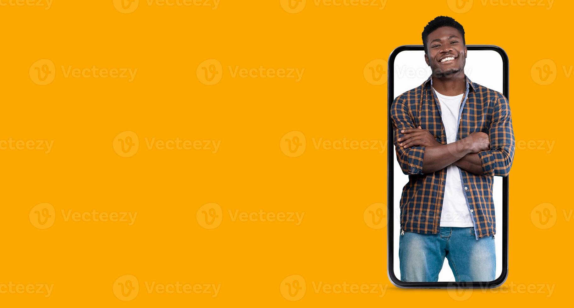 A person stands confidently with crossed arms, smiling at the camera, while appearing to be engaged with a smartphone featuring a large screen. The background is bright and inviting. photo