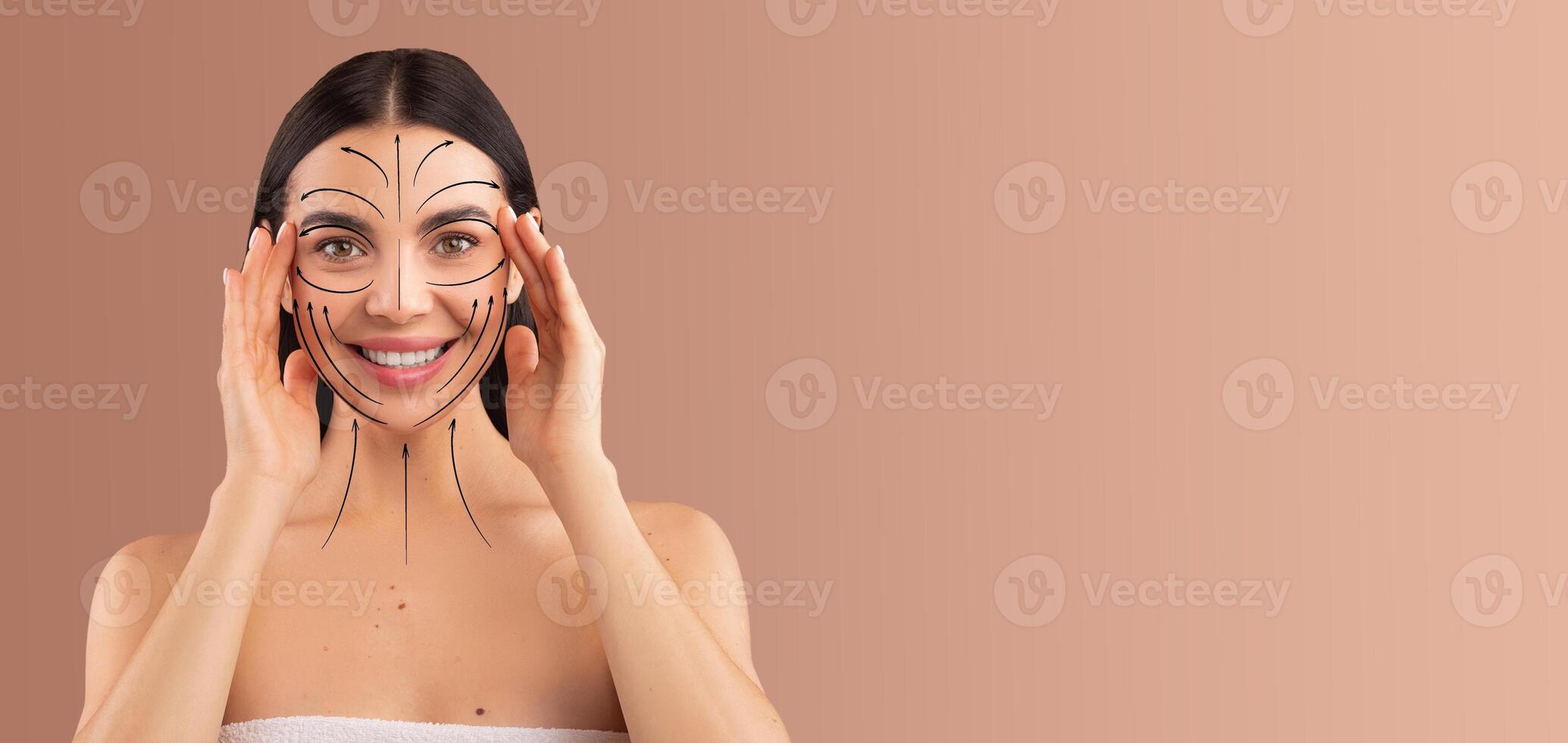 A woman is enjoying her beauty routine, using her hands to gently apply skin care techniques. She is isolated against a soft backdrop, emphasizing her joyful expression and commitment to self-care. photo