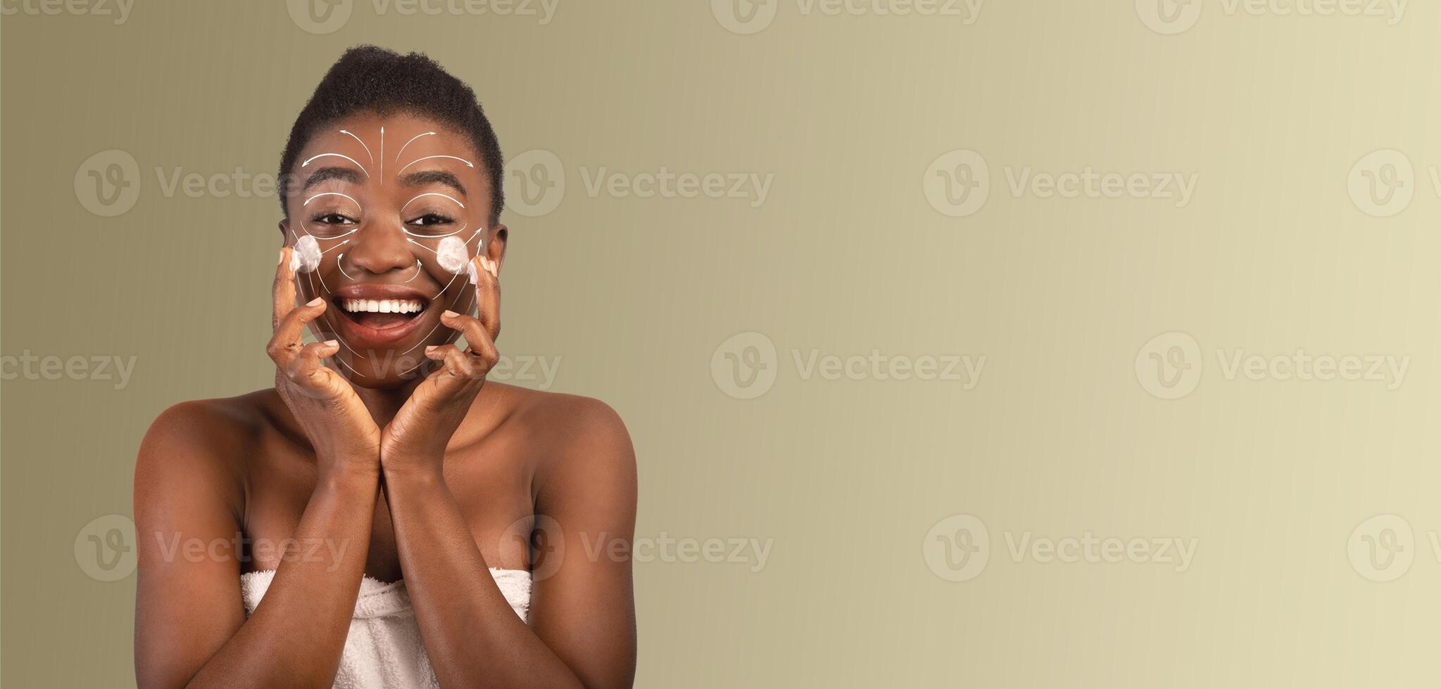 African American lady with a radiant smile applies a face mask while pampering her skin. She engages in her beauty routine, surrounded by a calming, neutral backdrop that enhances her joyful mood. photo