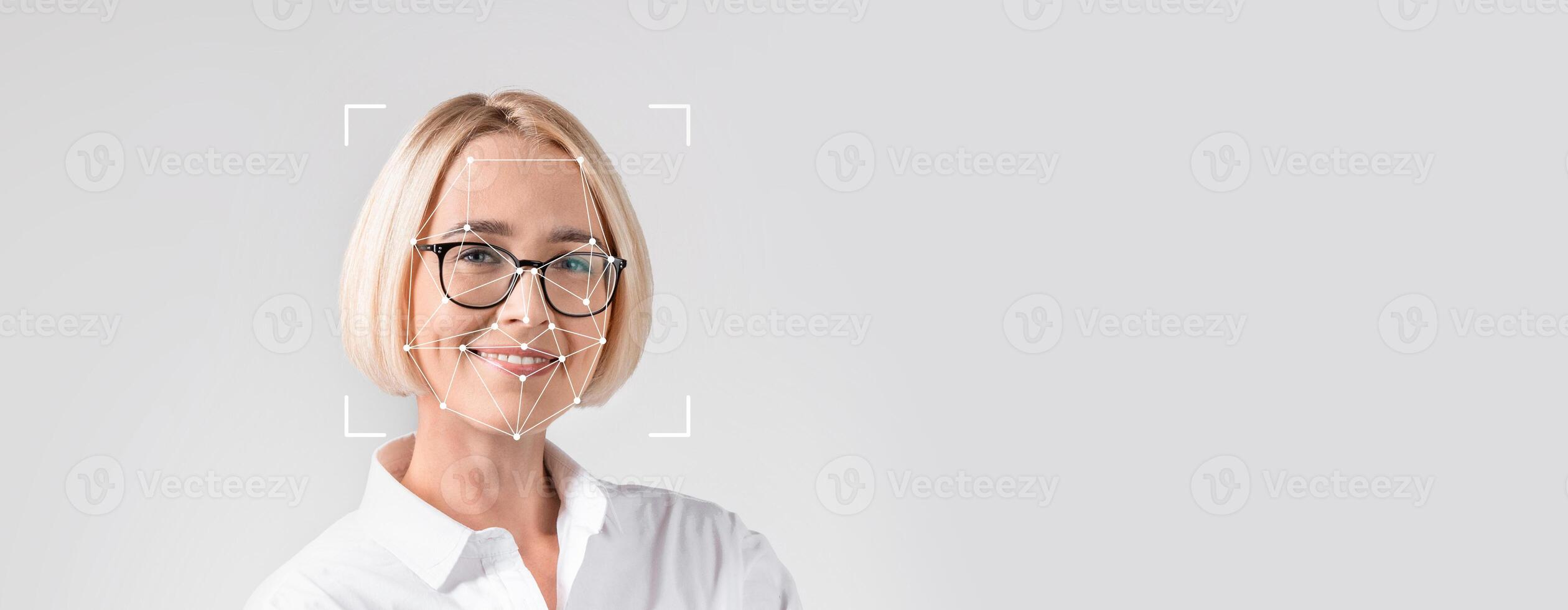A woman with short blonde hair and glasses stands alone in front of a blank background. Facial recognition overlays highlight her facial features for identification purposes. photo