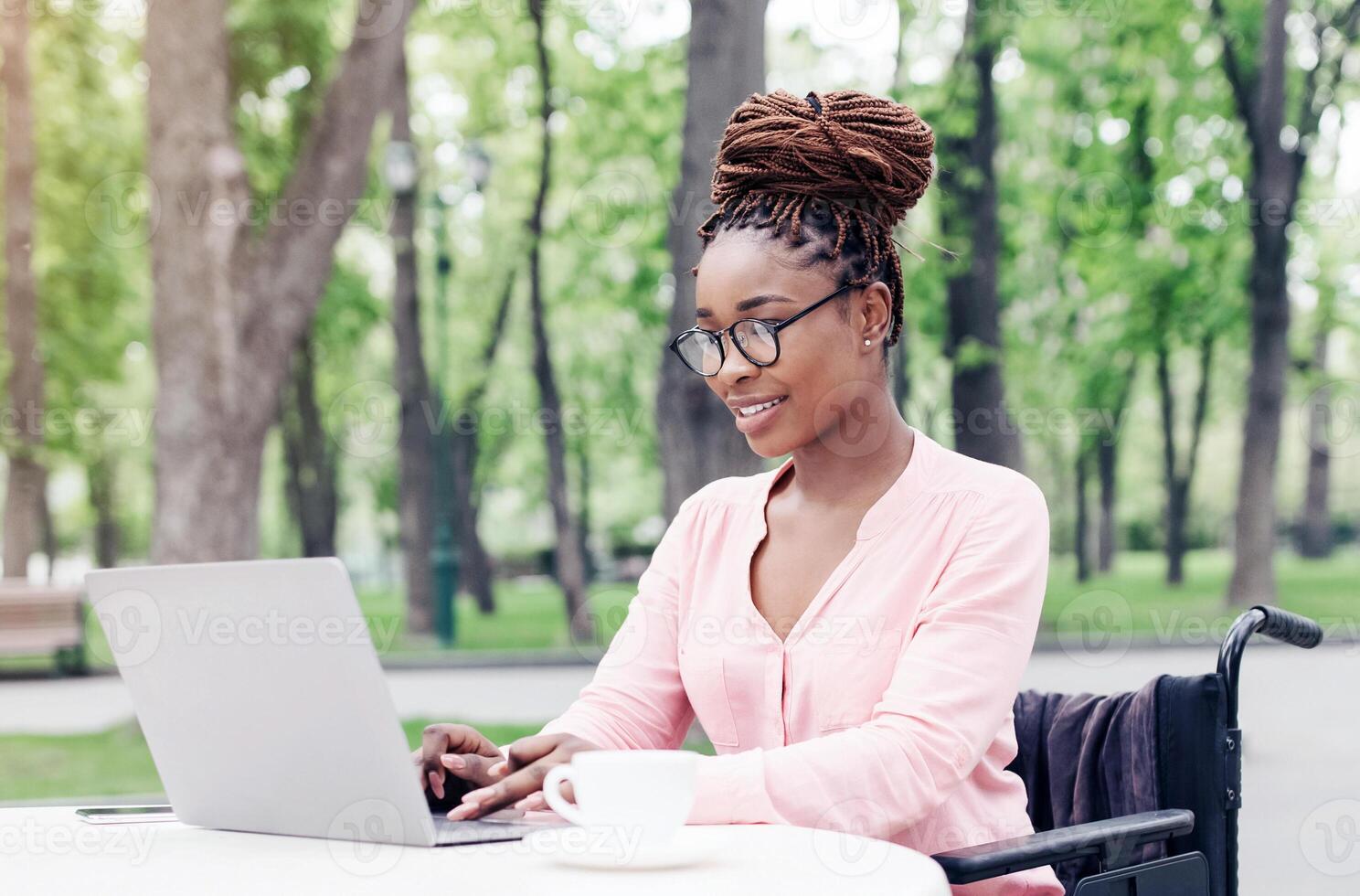 Positive young black woman in wheelchair using laptop in outdoor cafe at spring park, working on online business project, attending meeting remotely, having educational webinar, copy space photo
