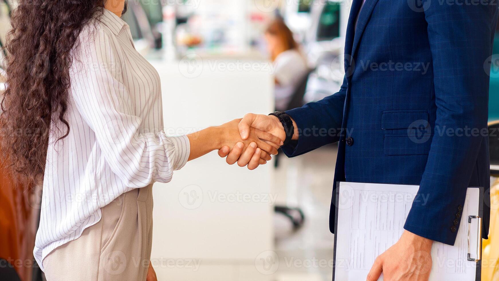 Cheerful sales assistant in formal outfit greeting pretty brunette lady customer, shaking hands and smiling at each other, huge salon of luxury automobiles interior. Curly woman buying new auto photo