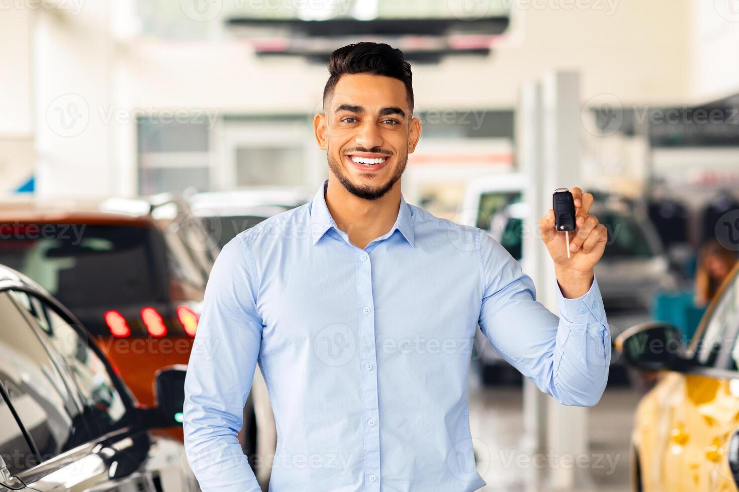 Happy muslim guy atnding among nice luxury and sports cars in huge auto showroom, showing key from automobile and smiling at camera, arab man got new vehicle at vip salon, copy space photo