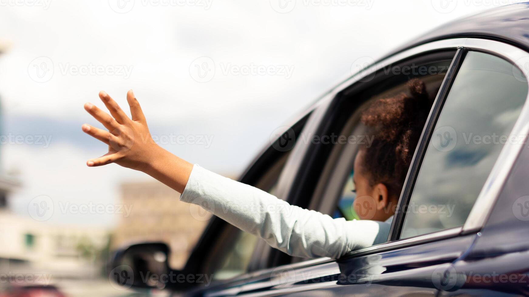 African American Kid Girl Putting Arm Out Of The Car Window Having Fun While Riding With Family In City. Summer Road Trip Adventure Concept. Back View, Selective Focus photo