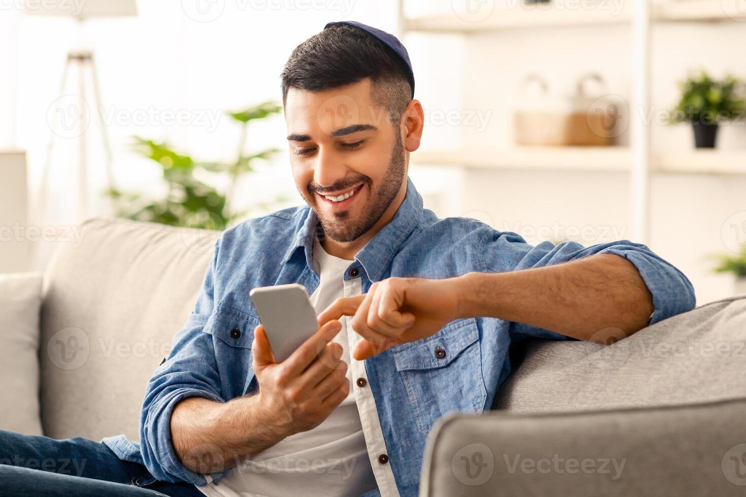 Useful App. Handsome Israeli Man In Kippah Using Smartphone While Relaxing On Couch At Home, Jewish Guy Sitting On Sofa In Living Room, Browsing Internet Or Messaging With Friends, Free Copy Space photo