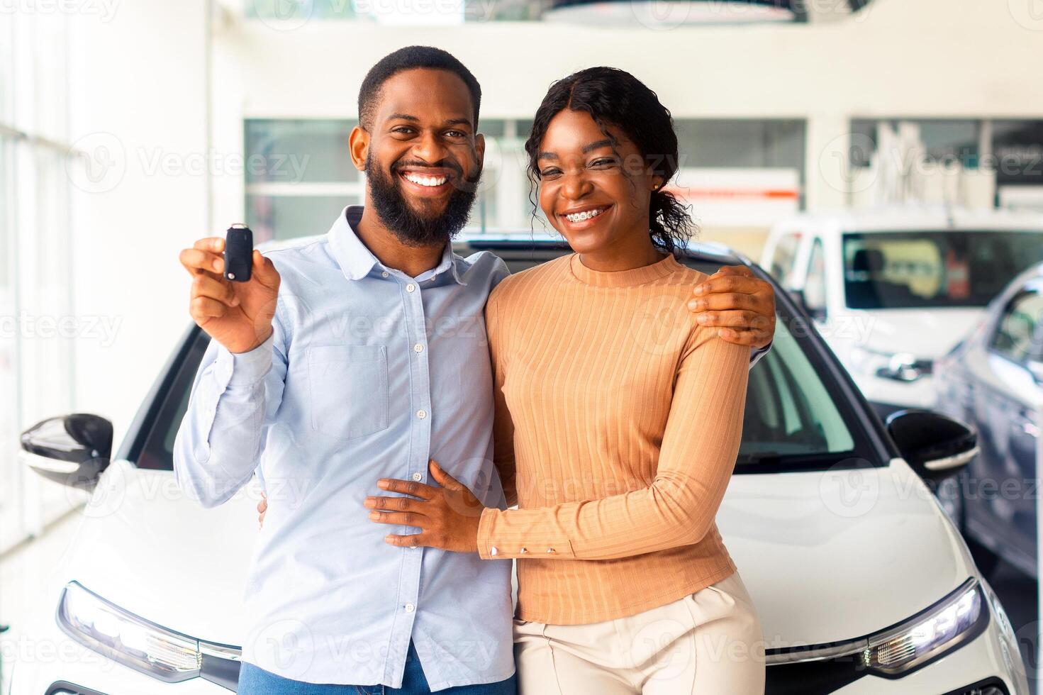 Happy Black Couple Posing With Car Keys In Hands In Modern Dealership Center, Cheerful African American Spouses Standing Near Their New Vehicle In Showroom And Smiling At Camera, Copy Space photo