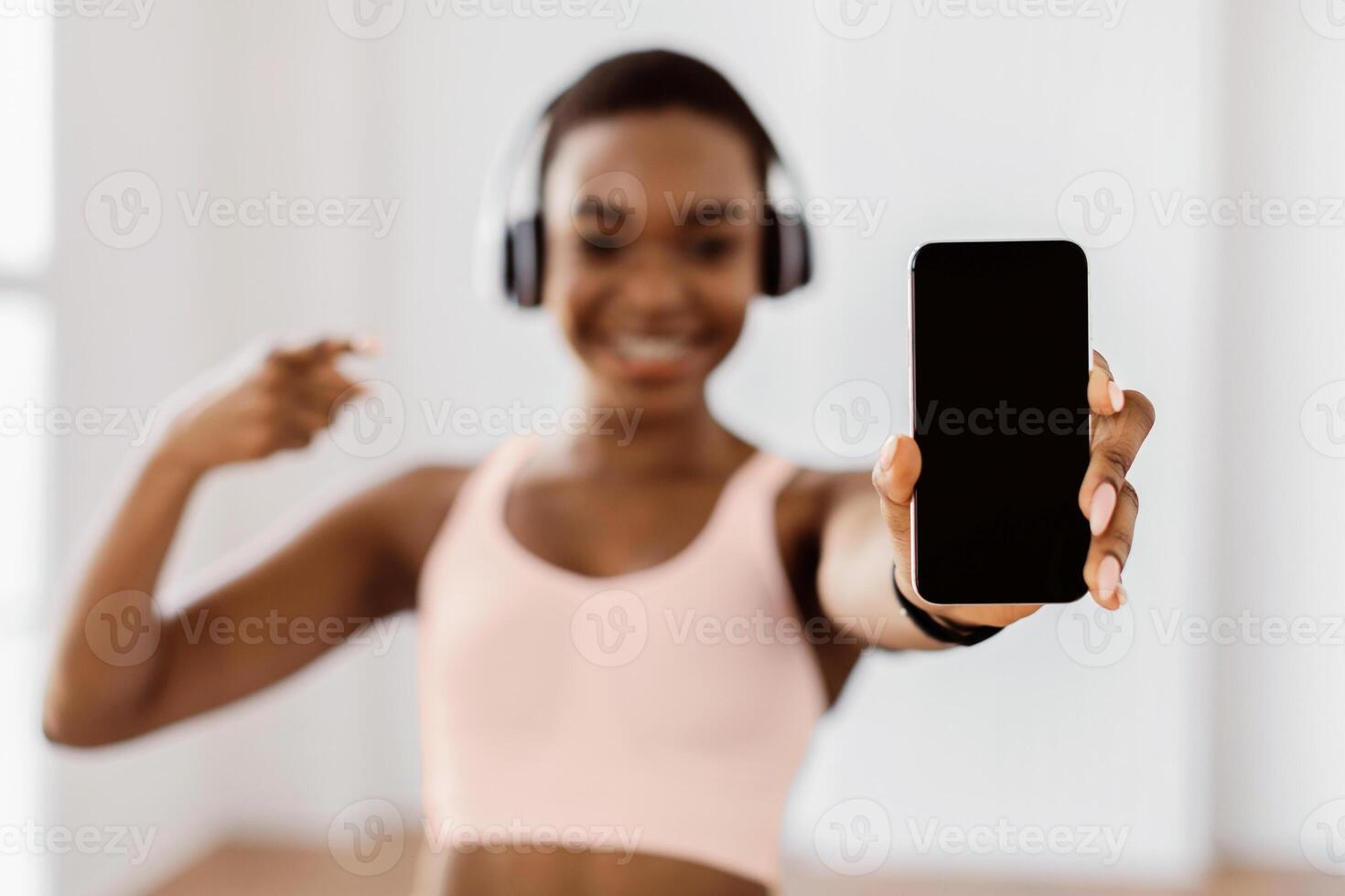 Fitness Application. Smiling slim young African American woman in sportswear holding mobile phone and pointing at isolated sporty empty screen for mock up template, selective focus, blurred background photo