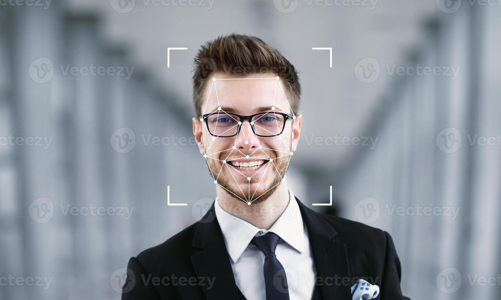 A young man with glasses and a confident smile stands in a modern setting. Facial recognition technology is applied to his face, highlighting its features against a blurred background. photo