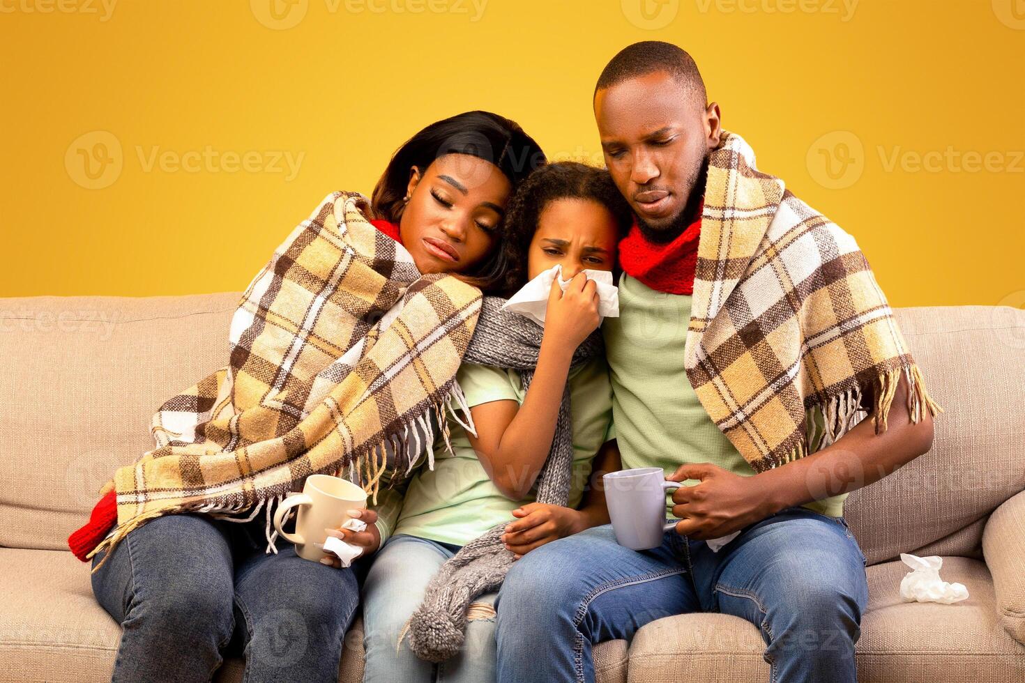 Flu season concept. Sick black parents and child girl feeling unwell, blowing runny noses and suffering from fever, sitting together on sofa over yellow studio background photo