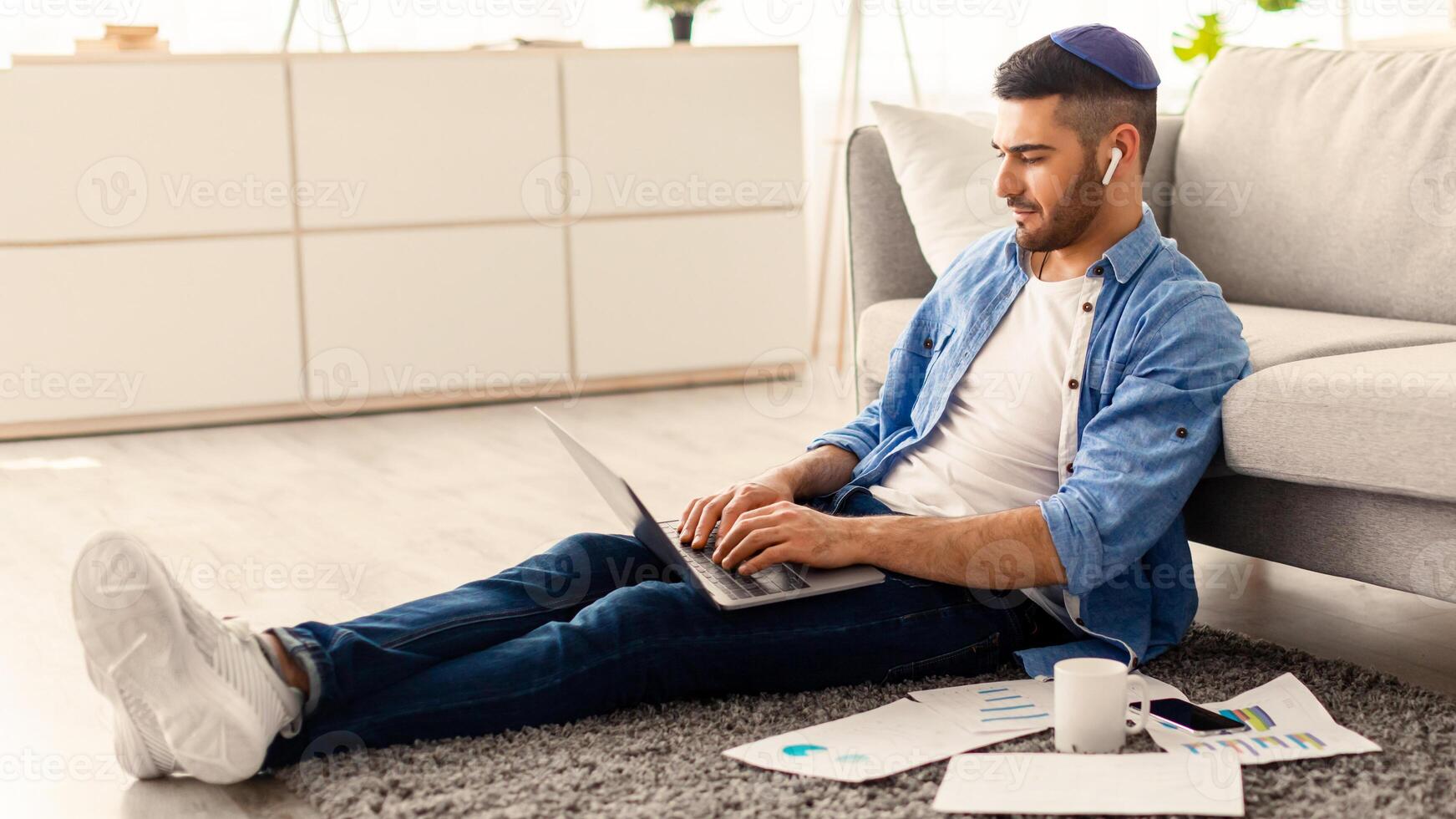 Serious millennial jewish man using laptop sitting on floor carpet leaning on couch, focused casual guy communicating online, writing emails, distantly working or studying on computer doing paperwork photo