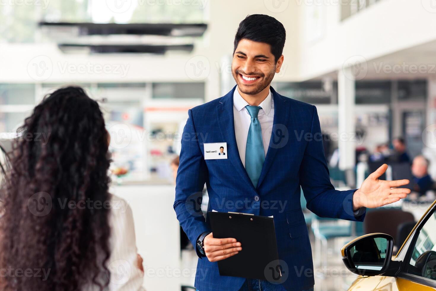 Wealthy middle-eastern young woman choosing new car, having conversation with friendly sales manager at brand new vip showroom, dealer pointing at yellow comfy auto and smiling, side view photo