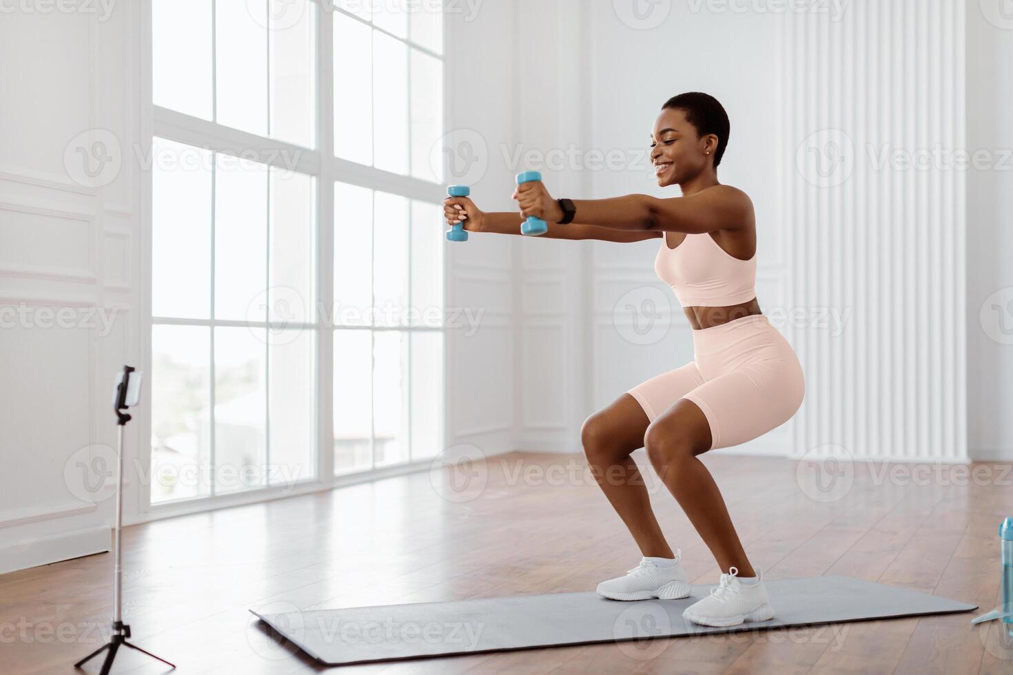Positive sporty lady exercising and squatting with two dumbbells in front of cellphone on tripod at home, smiling fitness blogger recording for her followers having personal virtual training photo