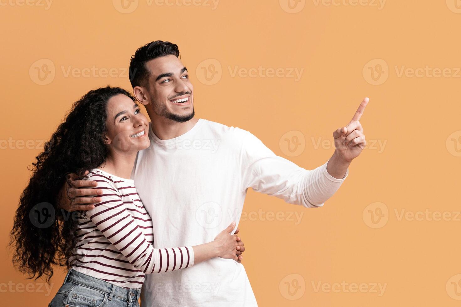 Nice Offer. Happy Young Arab Spouses Pointing Aside At Copy Space, Romantic Millennial Middle Eastern Couple Embracing And Showing Free Place For Your Advertisement Over Yellow Background, Panorama photo