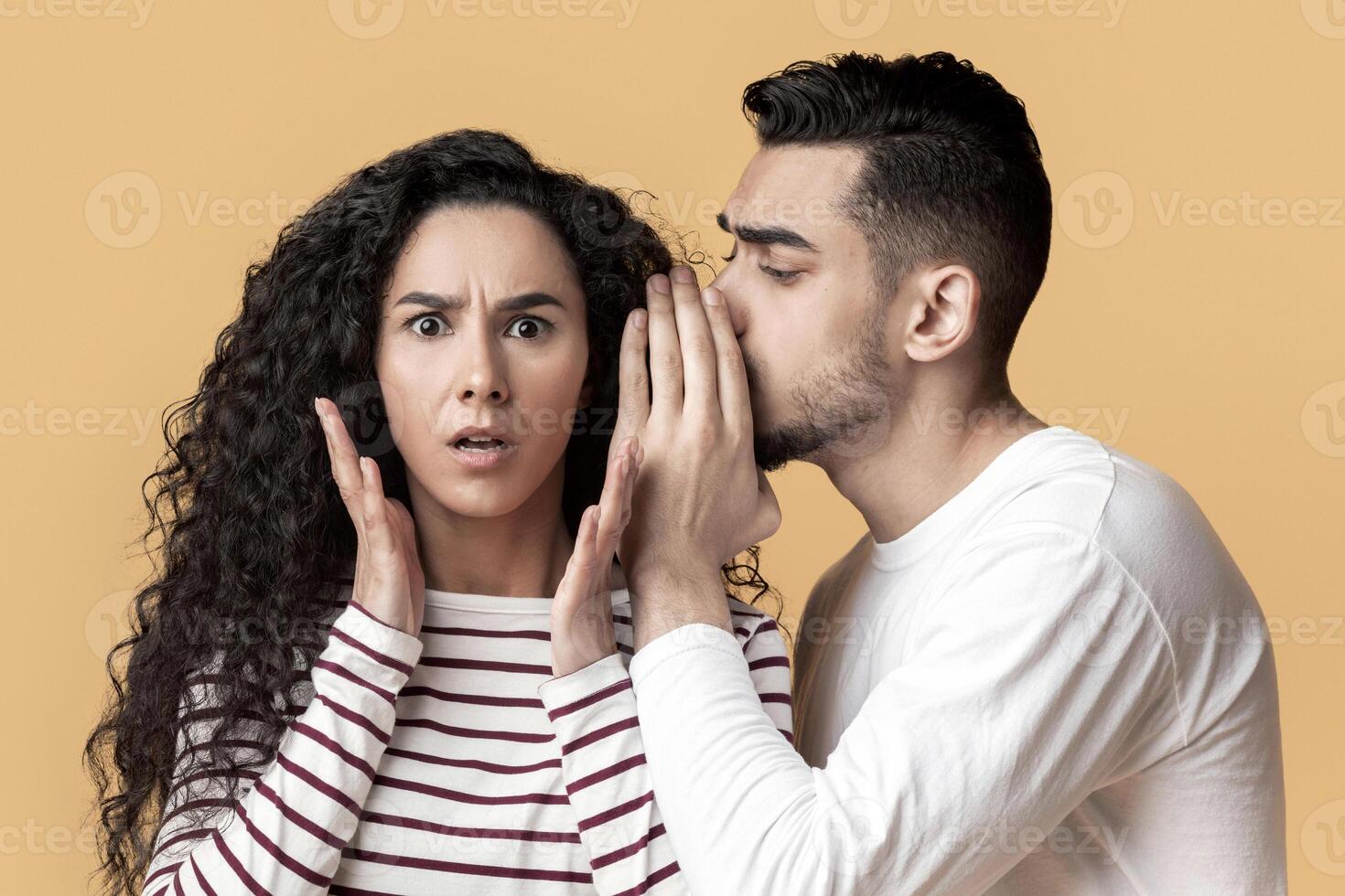 Big Secret. Young Arab Man Sharing Information With His Shocked Girlfriend, Whispering Something To Her Ear, Middle Eastern Guy Gossiping With Female Friend While Standing Over Yellow Background photo