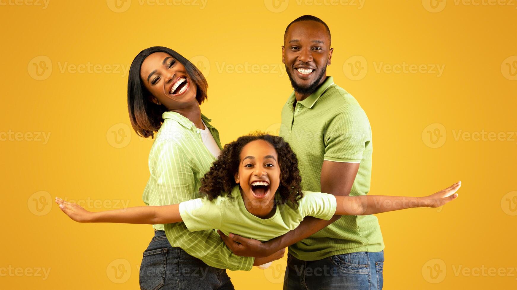 Family bonding concept. Joyful black man and woman holding daughter on hands, having fun over yellow studio wall. Smiling girl pretending she is flying, spreading hands, imitating plane photo