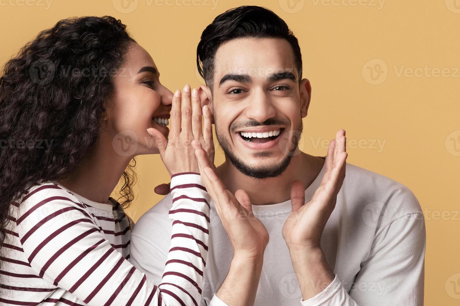 Gossips Concept. Lady Sharing Secret With Her Excited Arab Boyfriend, Joyful Middle Eastern Man Raising Hands In Amazement While Standing Together Over Yellow Studio Background, Empty Space photo