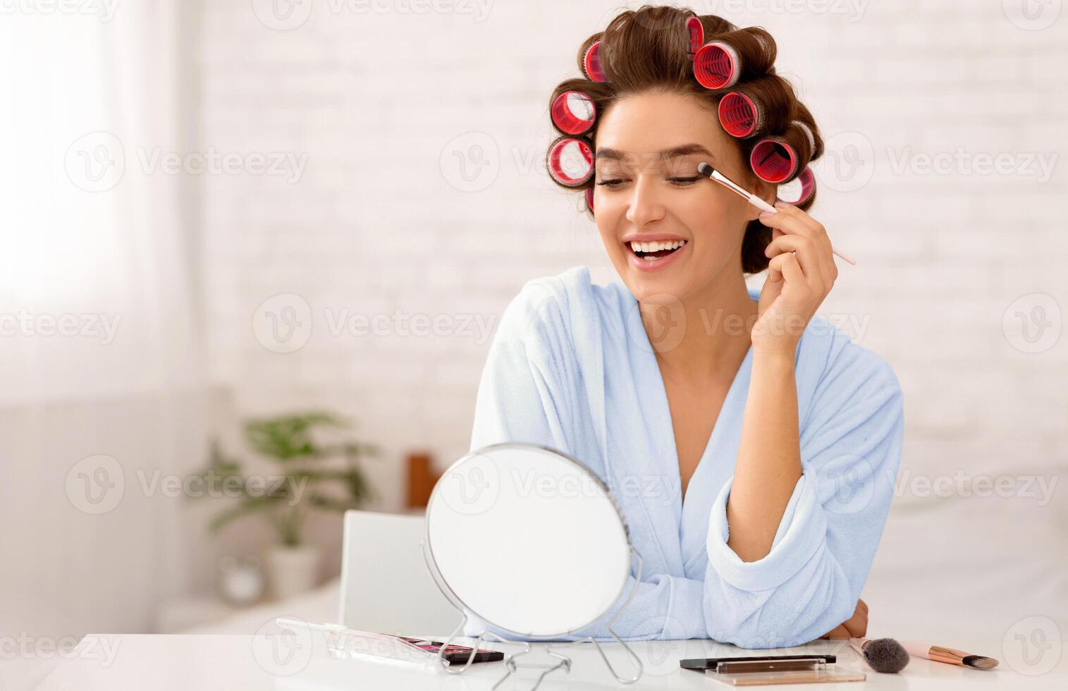 Glamorous Lady With Curlers Making Makeup Applying Shadow On Eye Lid Looking At Round Mirror Sitting In Modern Bedroom At Home. Female Beauty Routine And Cosmetics Concept. Selective Focus photo