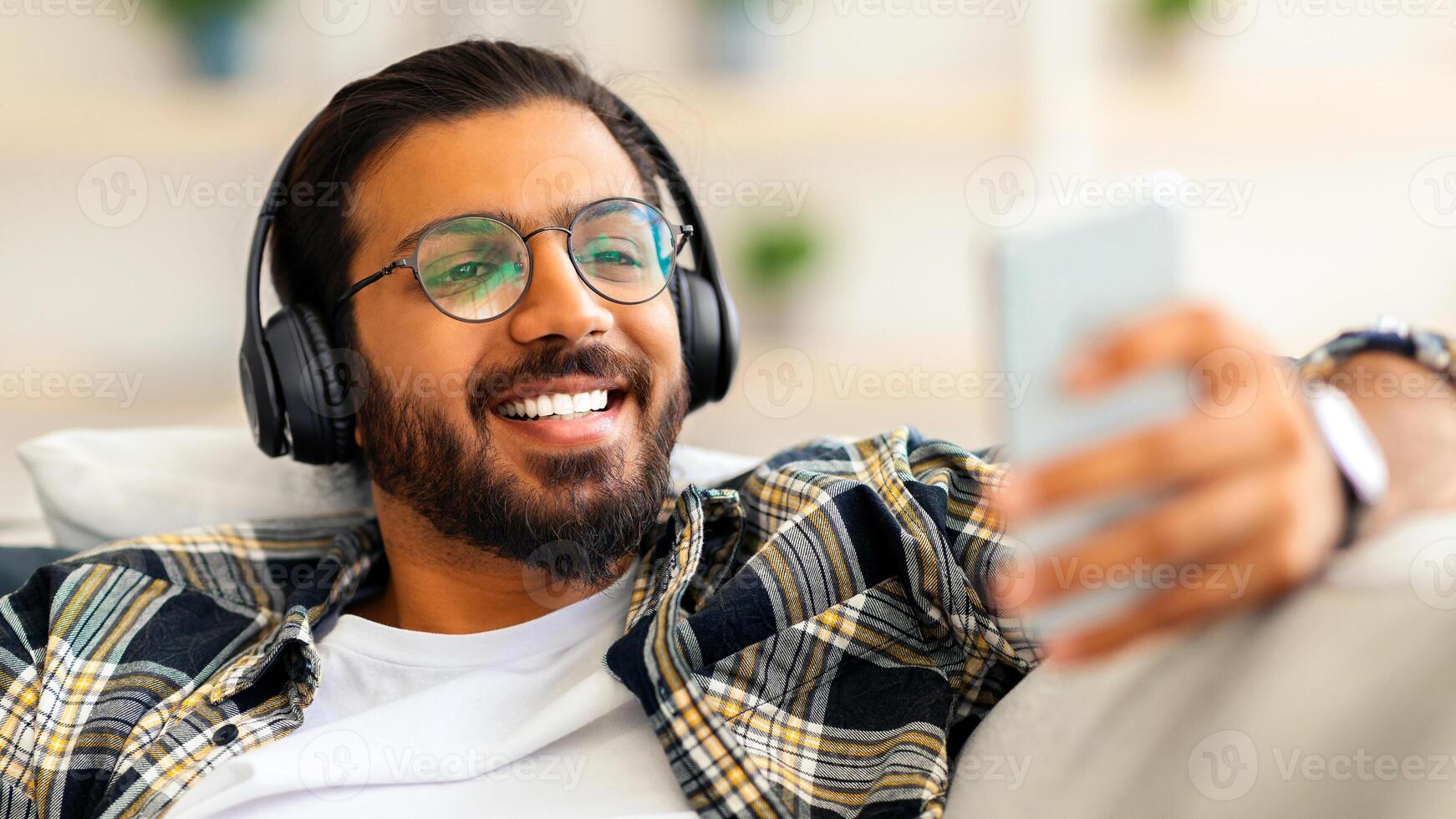 Handsome arab guy watching movie while resting on couch at home, using wireless headset and mobile phone, closeup. Joyful middle-eastern young man freelancer relaxing on weekend or having break photo