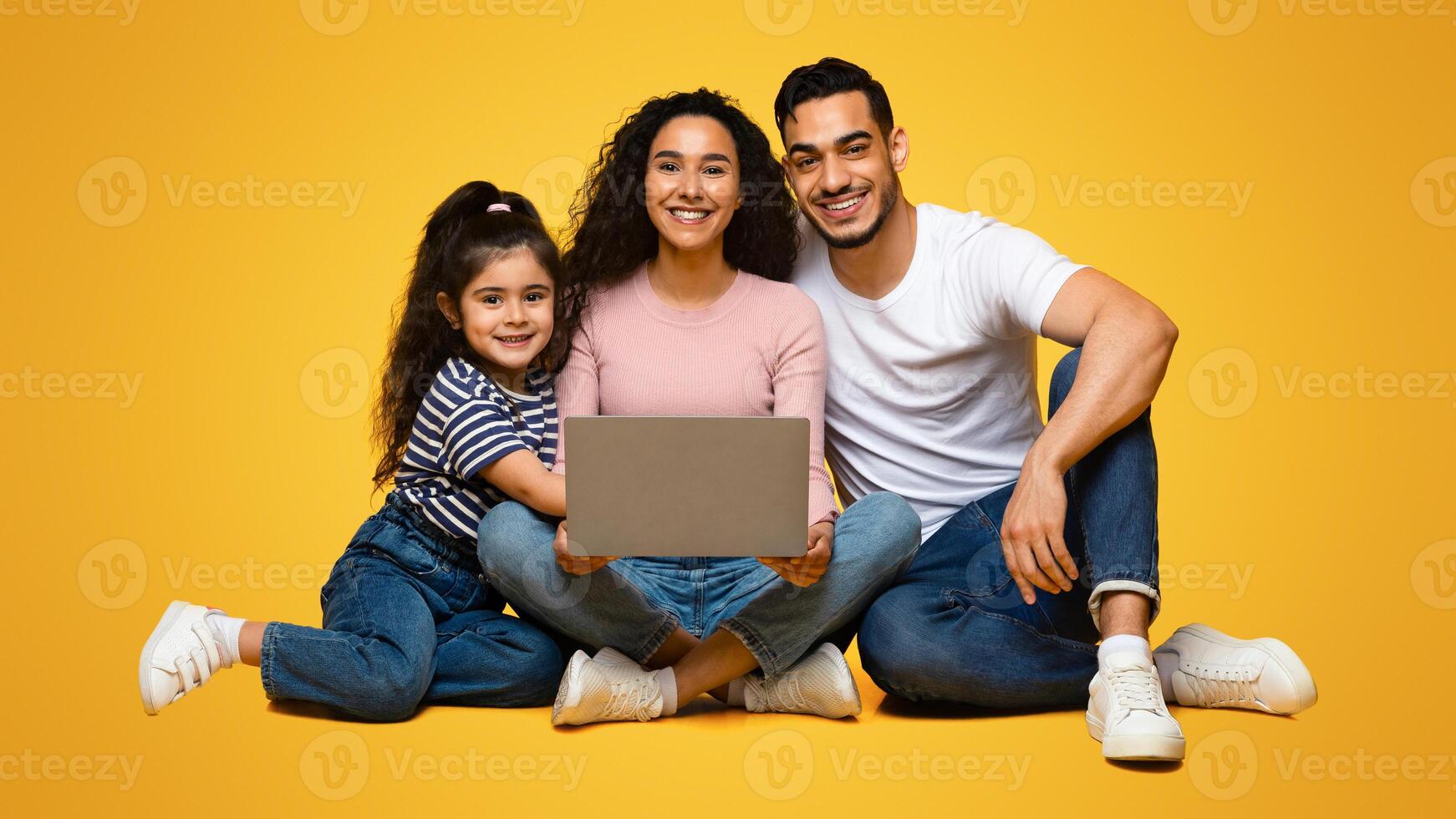Portrait Of Happy Beautiful Arab Family Of Three With Laptop Computer Posing Together Over Yellow Studio Background, Cheerful Middle Eastern Parents And Their Little Daughter Smiling At Camera photo