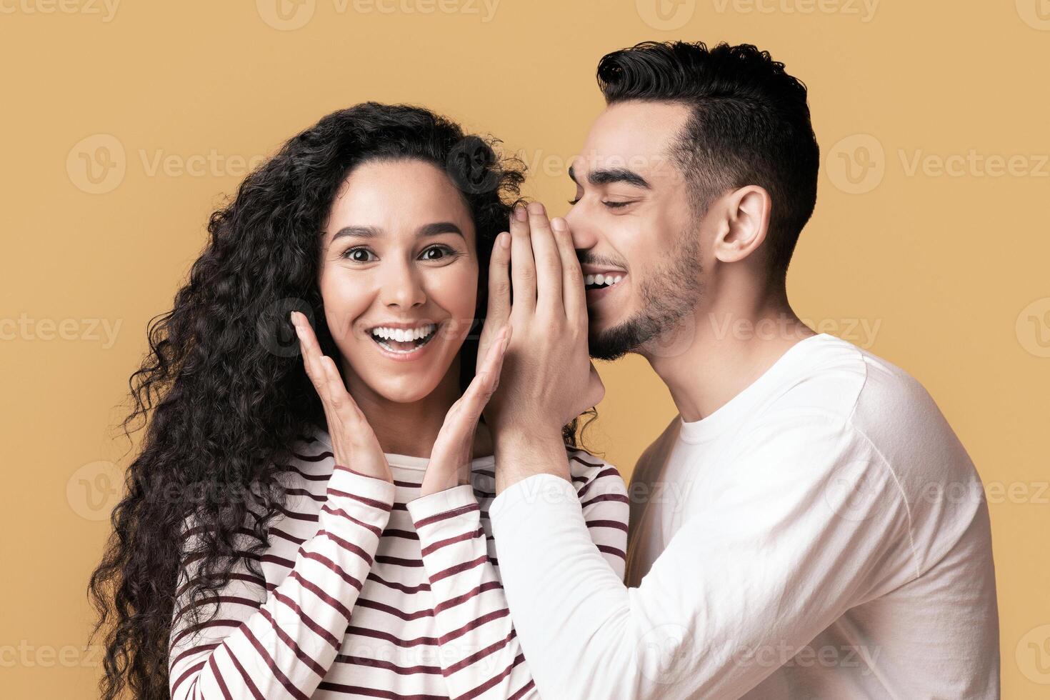 Big Secret. Young Arab Man Sharing News With His Excited Girlfriend, Surprised Middle Eastern Woman Raising Hands In Amazement While Standing Together Over Yellow Studio Background, Free Space photo