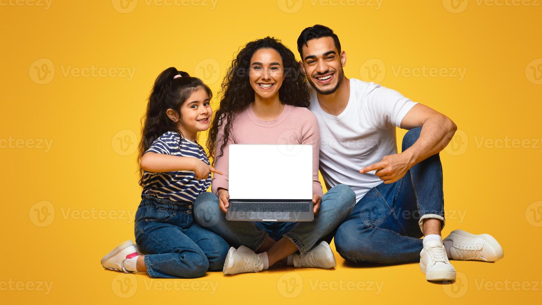 Cheerful arab family with little daughter showing laptop with blank white empty screen and pointing on it, happy middle eastern parents and female child recommending new website, mockup image photo