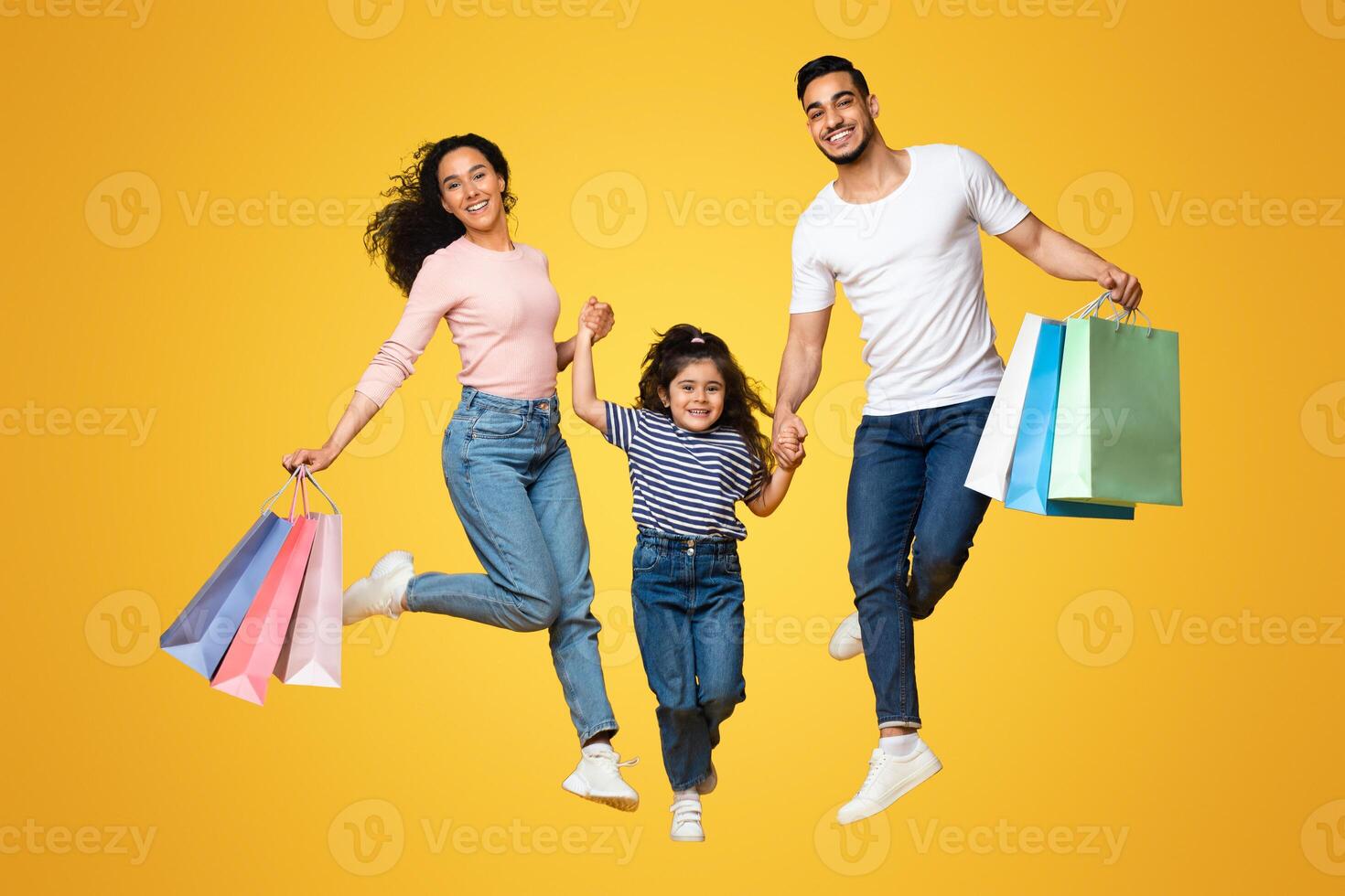 Big Shopping Concept. Overjoyed Arab Family Of Three Jumping With Lots Of Shopper Bags Over Yellow Studio Background, Middle Eastern Parents And Little Daughter Enjoying Seasonal Sales, Full Length photo