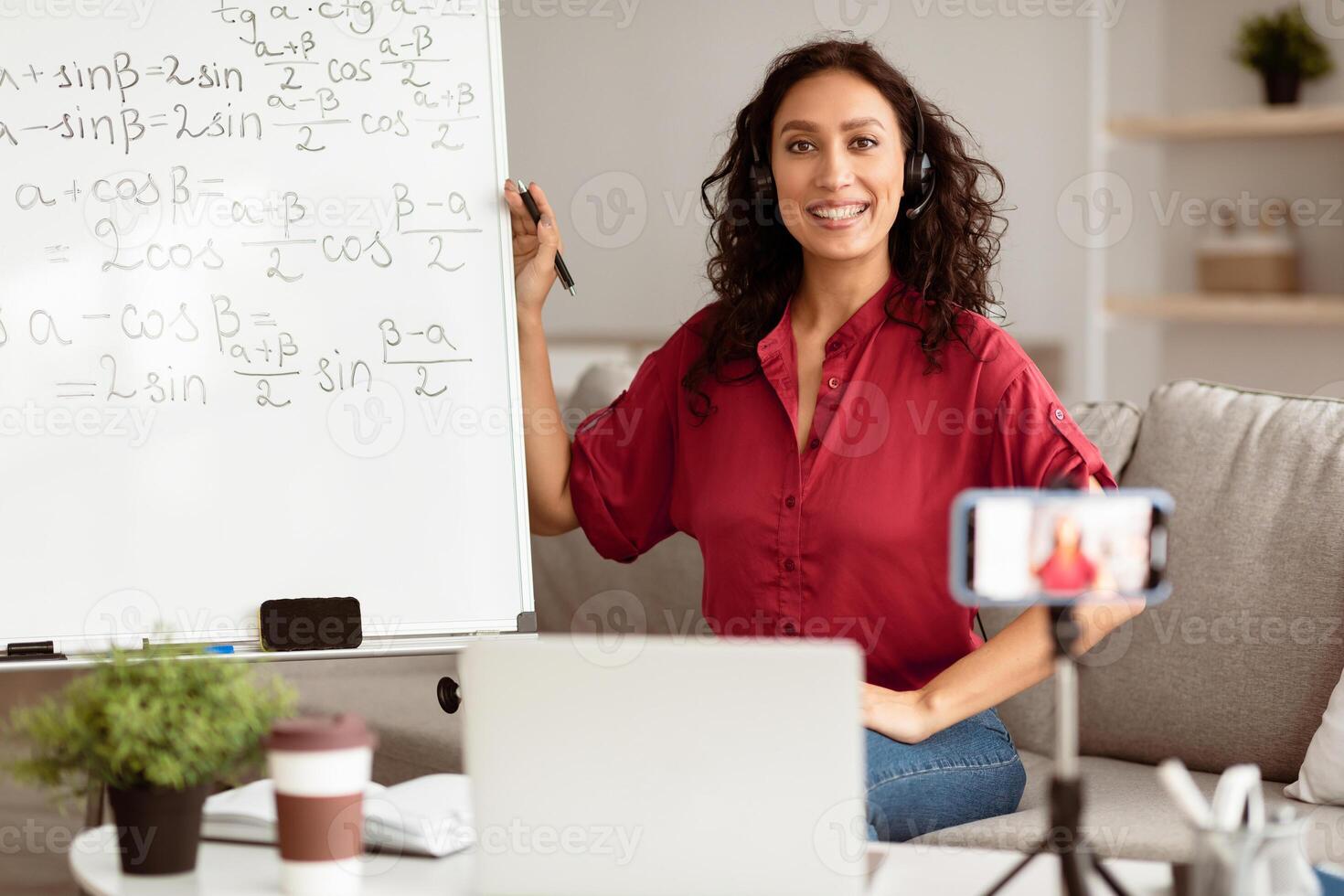 Online Learning Concept. Portrait of smiling lady teacher in headset sitting near whiteboard, explaining math formulas to students. Excited woman posing and looking at camera, recording class photo