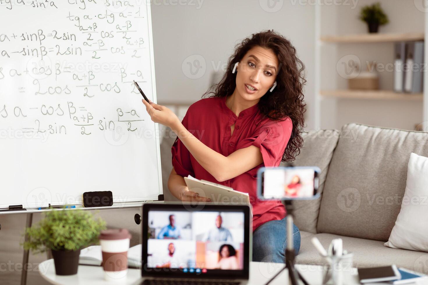 Online Lesson Concept. Portrait of young tutor pointing at board with trigonometric functions, explaining math to students making videochat. Woman in wireless headphones filming class on smartphone photo