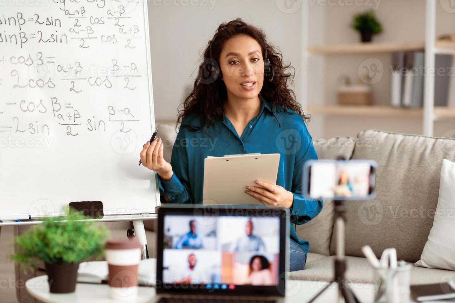 Modern Remote Education. Young woman having videochat with students, explaining mathematics formulas to her students. Teacher in wireless headset shooting training on smartphone using tripod photo