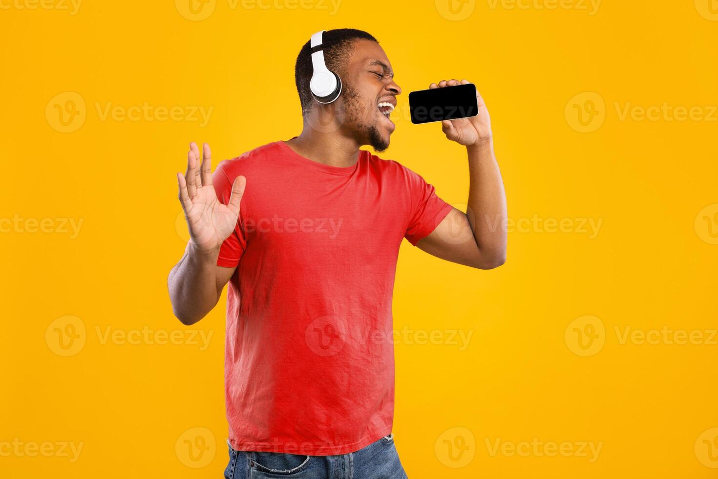 Black Man Singing Holding Smartphone Like Microphone Using Karaoke App Having Fun Standing On Yellow Studio Background. Guy Wearing Wireless Headphones Enjoying Music Online Using Application photo