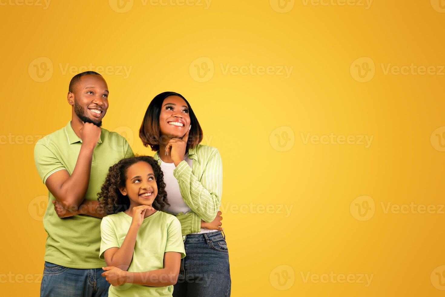 Special offer. Young black family of three looking aside at free space and holding hands on chin, standing over yellow background, studio shot, banner photo