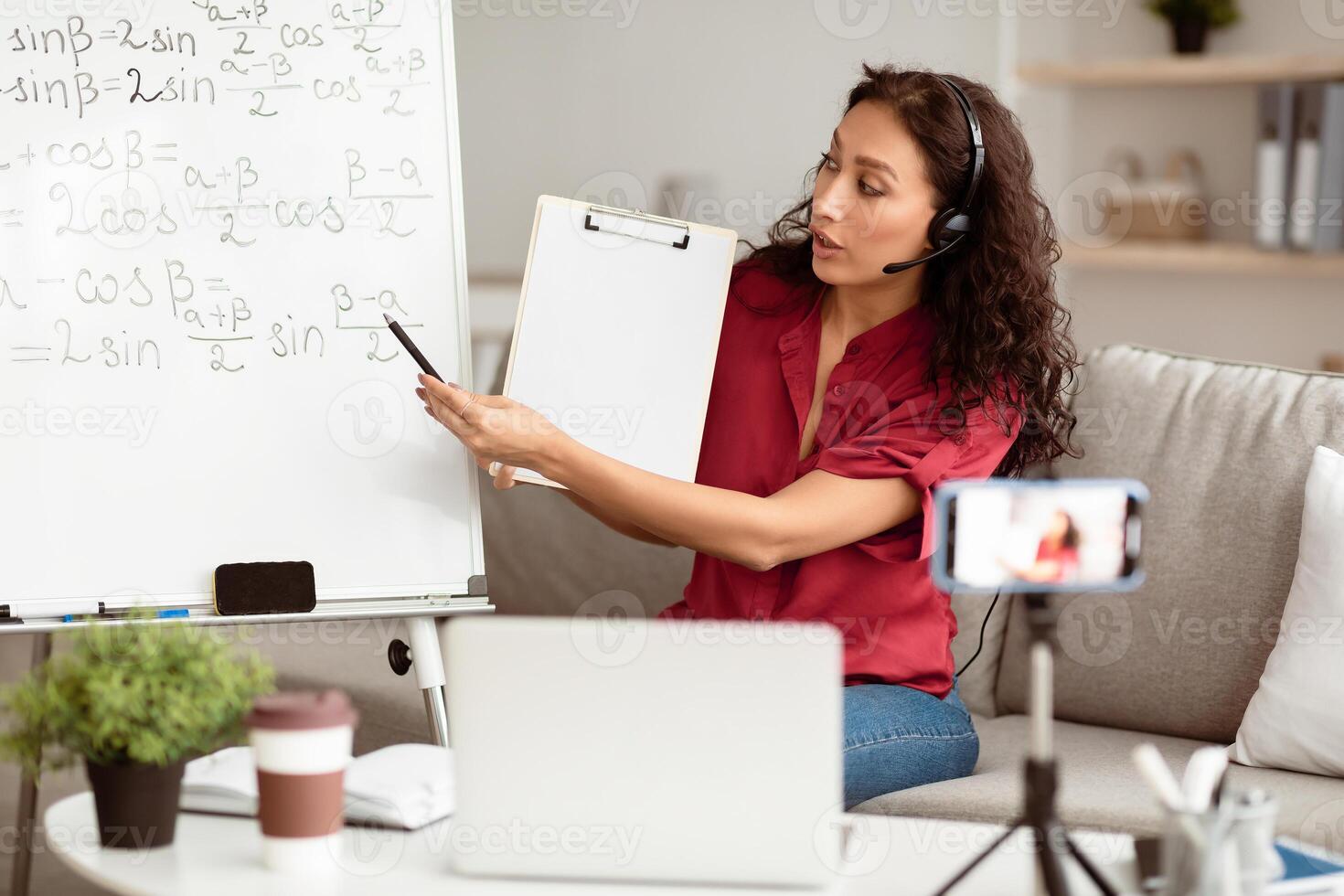 E-Learning. Confident woman having conference call using laptop. Lady teacher in headset showing clipboard, recording tutorial with mobile phone on tripod, streaming live pointing at board photo