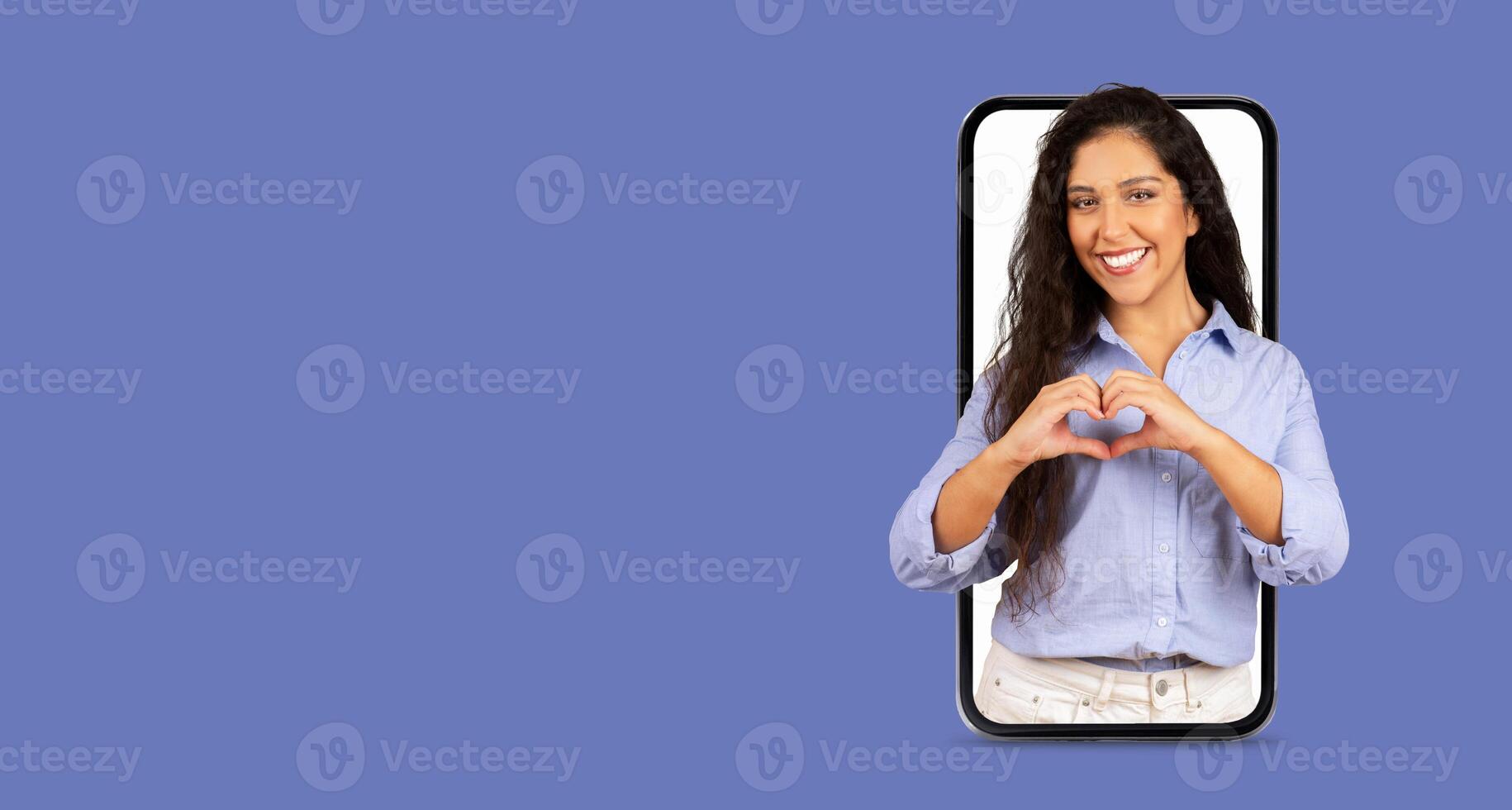 A young woman smiles while making a heart shape with her hands, showcasing her connection to online communication. The smartphone displays her image against a simple background. photo