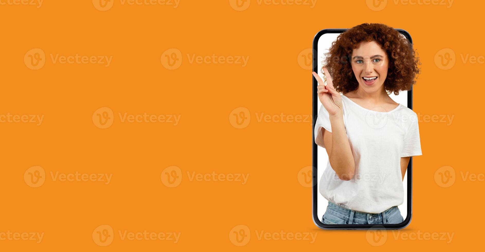 A cheerful individual with curly hair is making a peace sign while emerging from a large smartphone screen. The bright orange background enhances the playful mood and emphasizes technology. photo