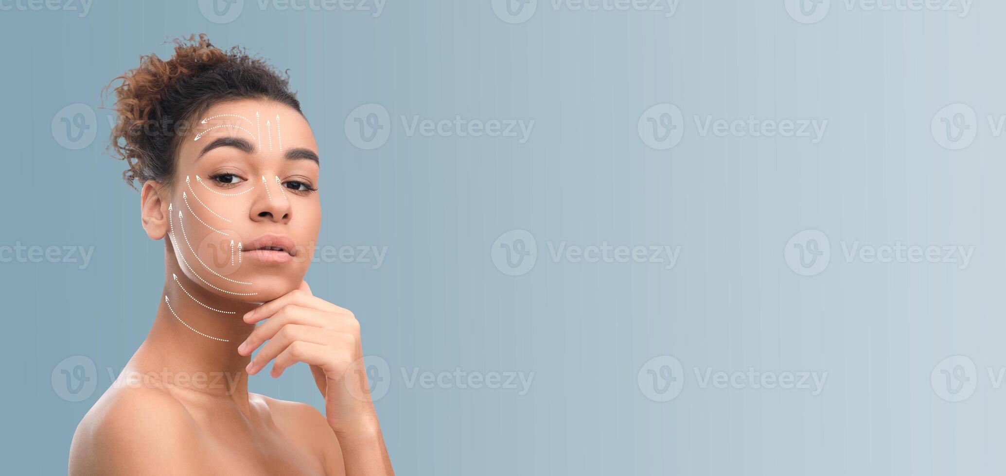 African American woman with curly hair is engaged in her skincare routine, gently touching her chin while displaying a calm demeanor. The background features neutral tones photo