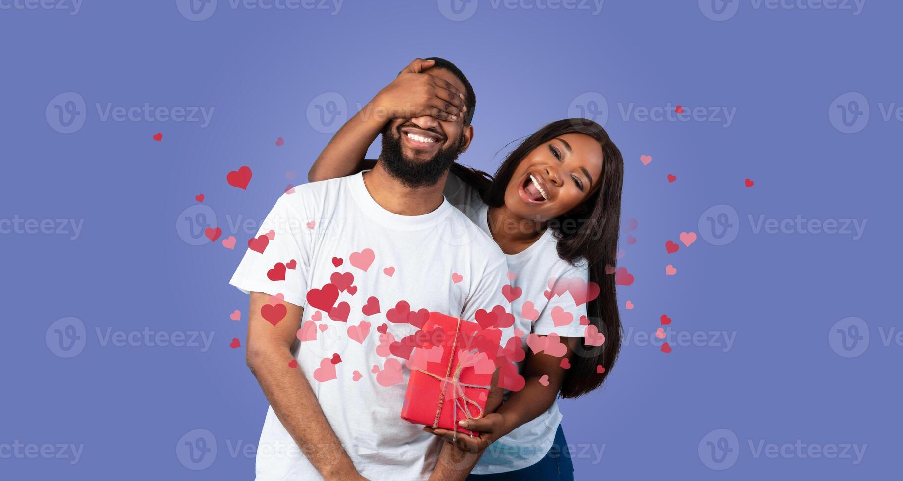 A happy black couple shares a loving kiss while surrounded by floating hearts. They celebrate their special bond with a red gift, embodying affection and joy on Valentine's Day. photo