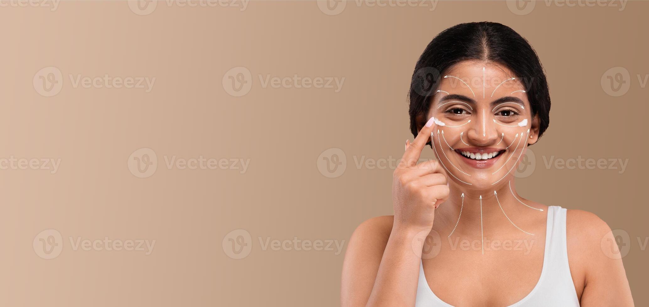 Indian woman engaged in her skincare routine smiles brightly as she applies a product to her face. The bright and minimal background highlights her joyful expression and self-care moment. photo