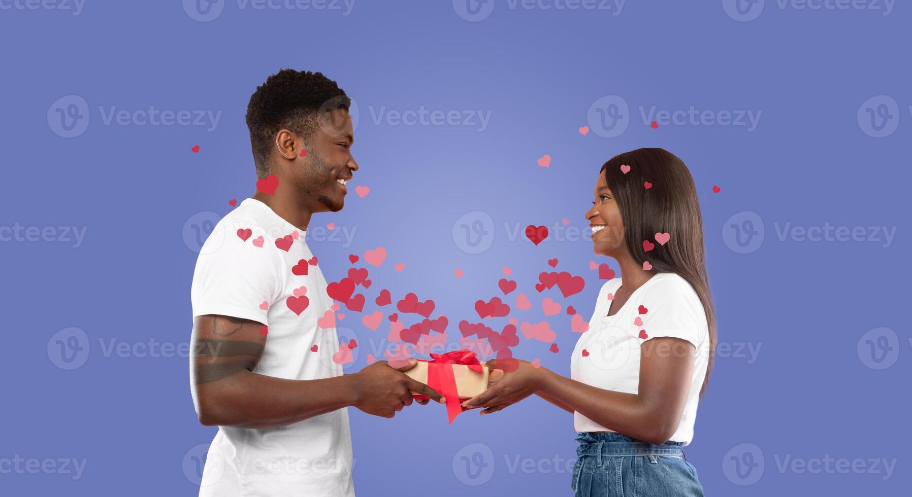 A black couple shares a joyful moment, exchanging a gift while surrounded by floating hearts. Their smiles reflect love and affection, making this a memorable celebration of Valentine's Day. photo