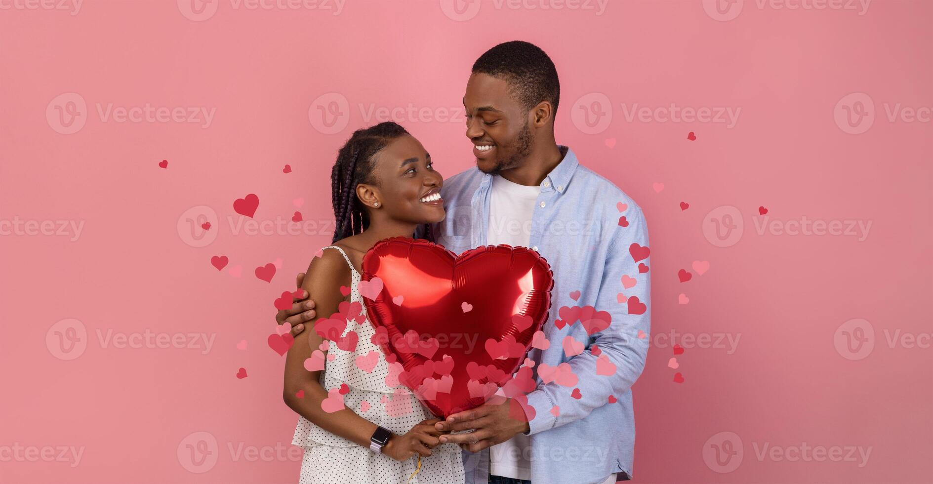 Black couple joyfully celebrates their love, sharing a kiss while holding a large red heart balloon. They are surrounded by floating hearts, symbolizing their bond and affection. photo