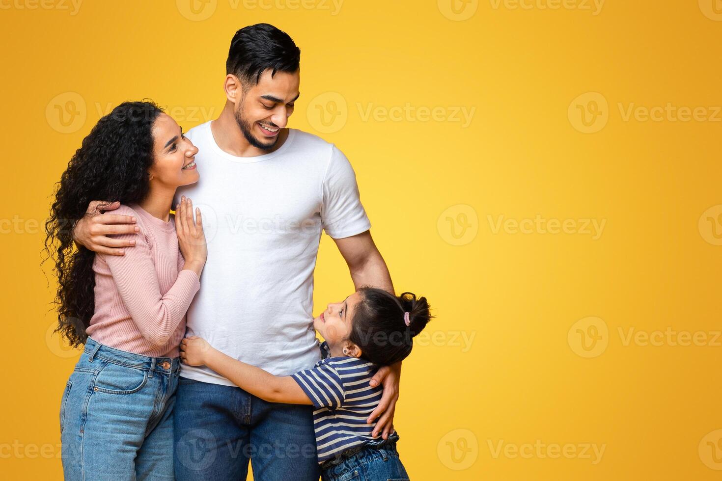 Full Length Shot Of Happy Middle Eastern Family Of Three Standing Isolated Over Yellow Background, Loving Arab Parents And Their Little Daughter Embracing And Smiling To Each Other, Copy Space photo