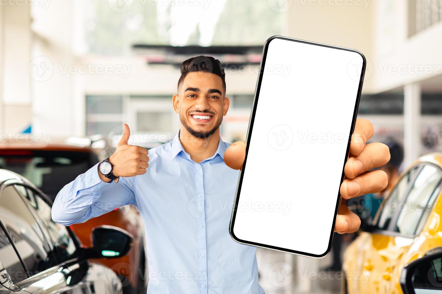 Handsome arab guy showing smartphone with white blank screen and thumb up, using mobile application while choosing new car at automobile dealership, reading recommends online, mockup photo
