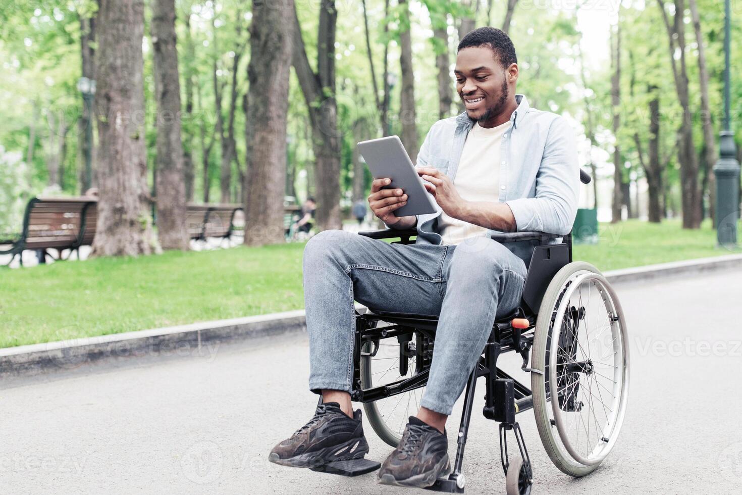 Positive young black guy in wheelchair using tablet computer, browsing web or watching at urban park, copy space. Full length of African American man checking social media on touch pad outdoors photo