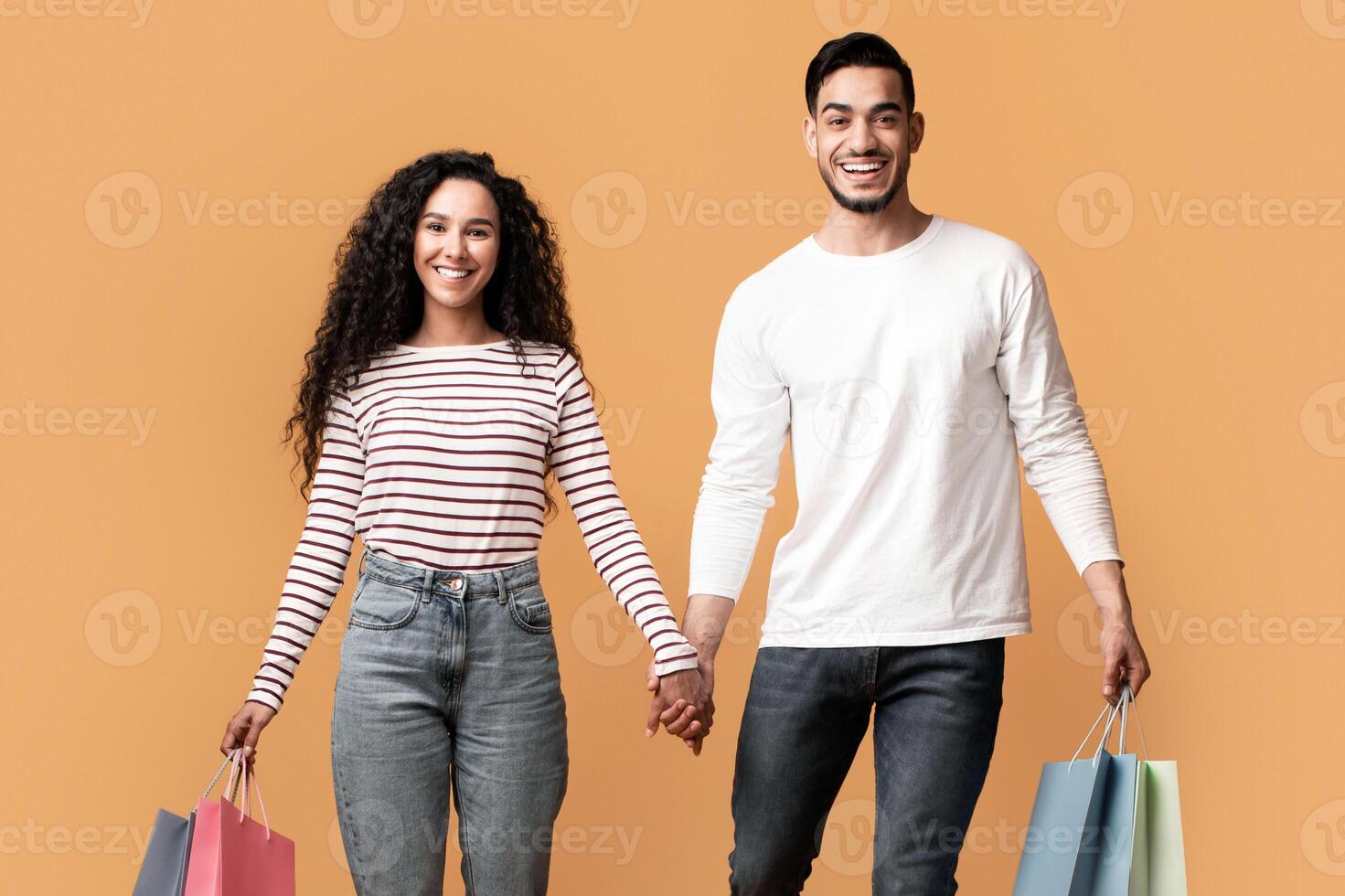 Portrait Of Happy Middle Eastern Couple Walking With Bright Shopping Bags Over Yellow Studio Background, Cheerful Arab Man And Woman Holding Hands And Enjoying Making Purchases Together, Full Length photo