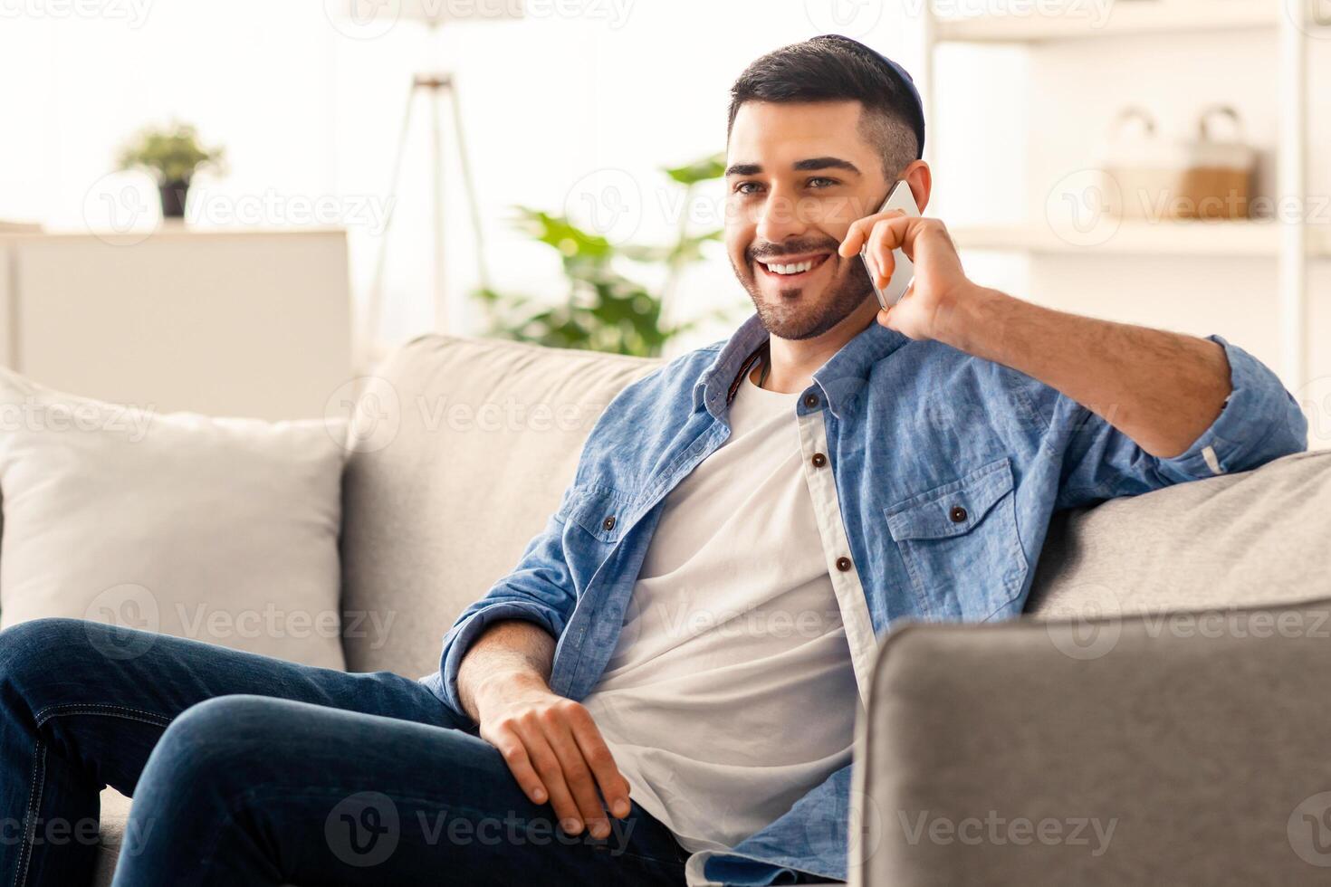 Happy cheerful young man talking on the phone at home, smiling jewish male making or answering call by cellphone sitting on sofa, handsome guy having pleasant conversation speaking by mobile photo