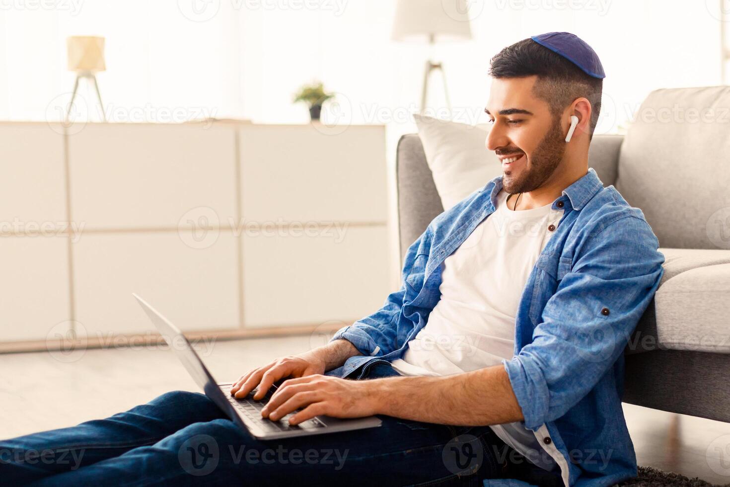 Handsome jewish freelancer guy working remotely with laptop at home office, using computer for online project or education, typing on keyboard, sitting on floor leaning on sofa, free copy space photo