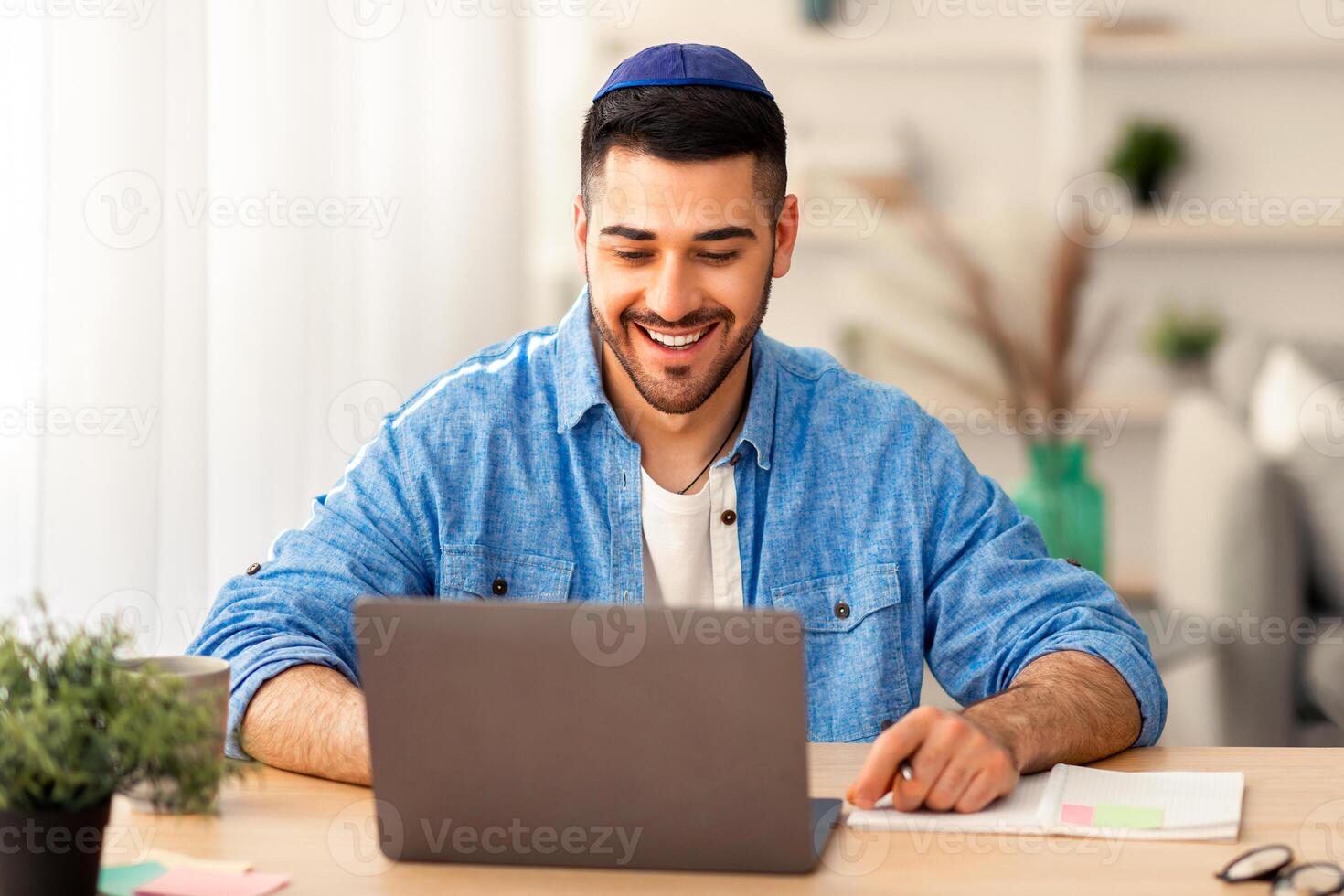 Telecommuting Concept. Potrait of smiling casual israeli man sitting at desk, using laptop. Male watching webinar looking at pc screen, working or studying remotely, free copy space, banner photo