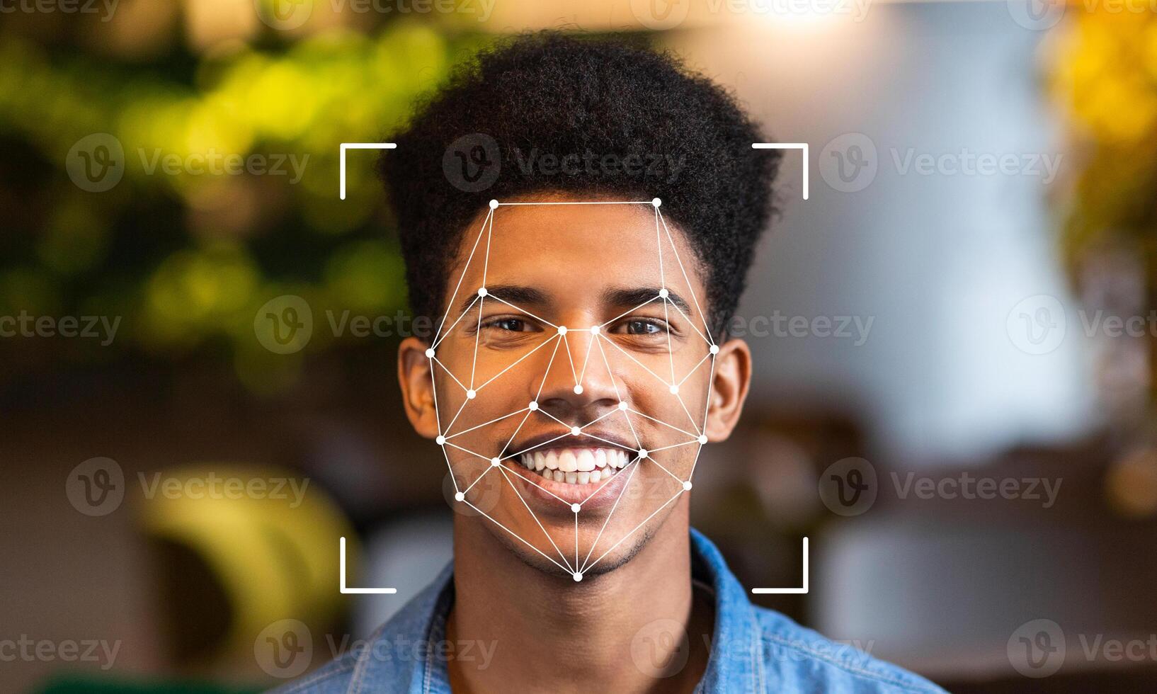 A young man is smiling while facial recognition technology scans his face. This advanced identification method is shown in a modern, well-lit environment with green foliage. photo