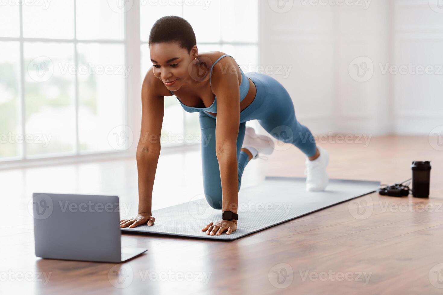 Online Training Concept. Sporty African American lady doing mountain climbers exercise, watching tutorial or having online class on computer, looking at screen, exercising in spatious apartment photo