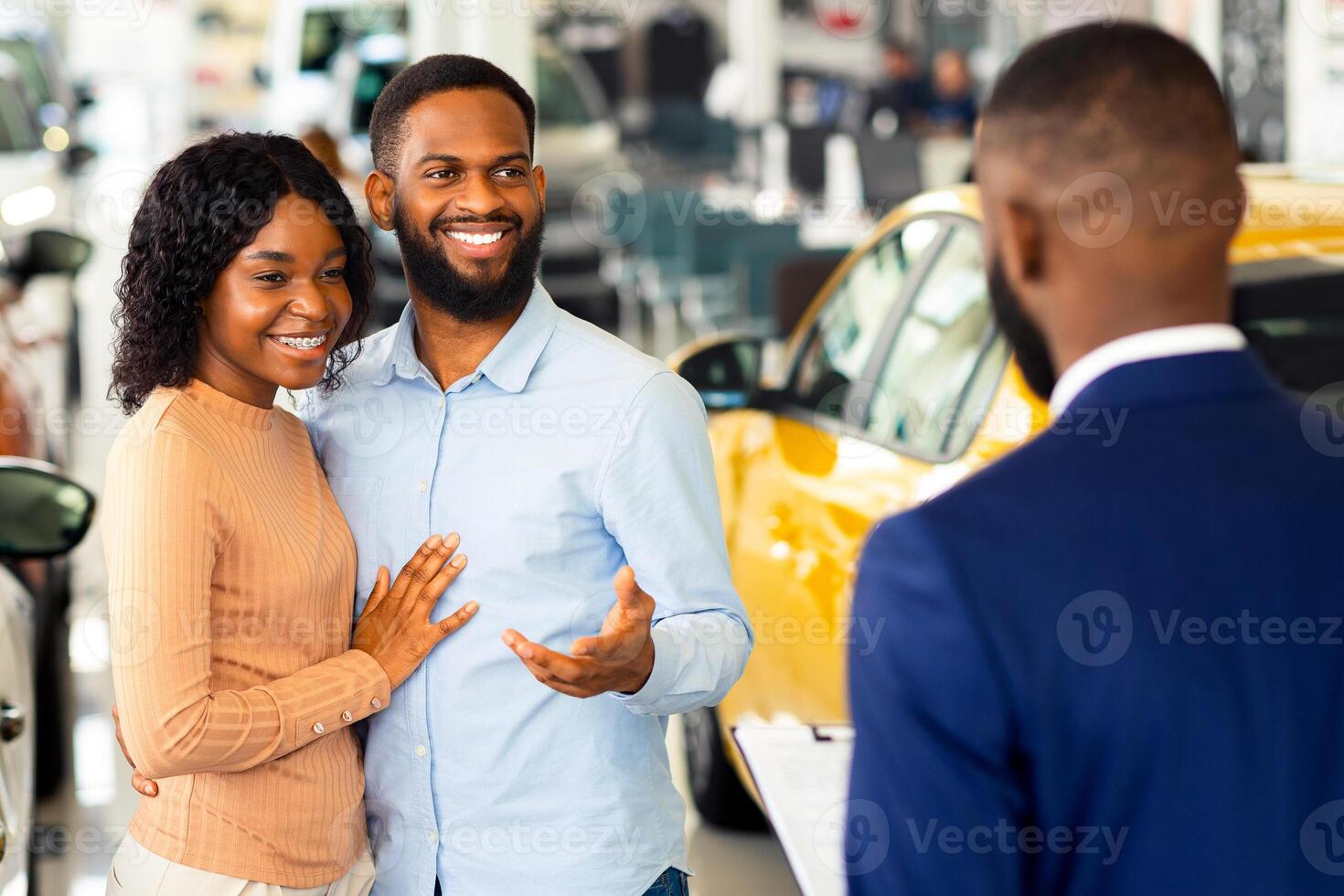 Young Black Couple Talking To Salesman In Dealership Office While Buying New Car, Happy African American Spouses Discussing Automobile Models With Salesperson In Showroom, Selective Focus photo