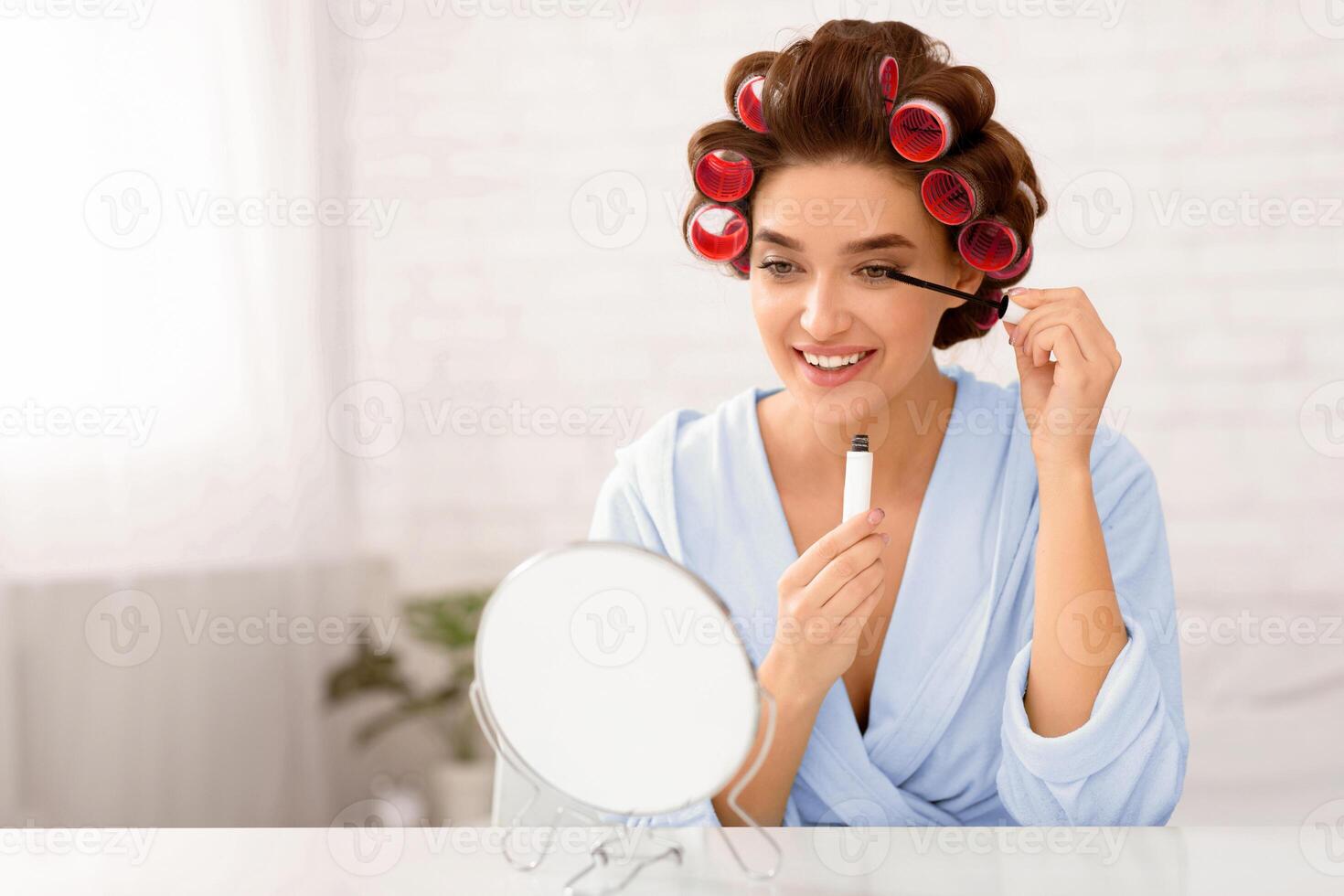 Makeup Cosmetics. Glamorous Woman With Curlers Applying Mascara On Eyelashes Looking At Round Mirror Sitting In Bedroom At Home. Female Beauty Routine Concept. Selective Focus photo